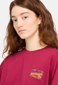 Camiseta de algodón burdeos con cuello redondo, que presenta un colorido parche del logo de Timberland en el pecho. Textura suave con mangas cortas.