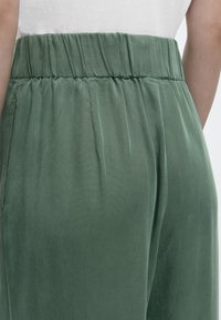 Πίσω όψη ατόμου που φοράει χαλαρά ελιάς πράσινα παντελόνια με ψηλή μέση και ελαστική μέση πάνω από λευκό τοπ.