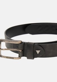 Cintura in pelle nera con una fibbia rettangolare in argento che presenta un'incisione "GUESS" e un accento triangolare sulla cinghia. Superficie testurizzata.