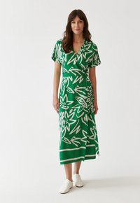 Grünes Wickelkleid mit weißem Blumenmuster, kurzen Ärmeln und einer gebundenen Taille. Getragen mit weißen Sneakers. Glatter Stoff, knielange Design.