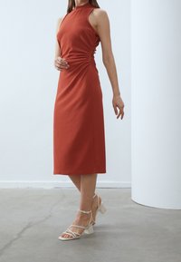 Šatové midi šaty v rezavé barvě s halter nákrčníkem, bočním ruching detailem a ribovanou strukturou; doplněné bílými sandály na blokovém podpatku se řemínky.