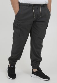 Dunkelgraue Cargo-Hosen mit elastischen Bündchen, seitlichen Taschen und einem verstellbaren Bund. Hergestellt aus einem weichen, strapazierfähigen Stoff. Kombiniert mit schwarzen Sneakers.