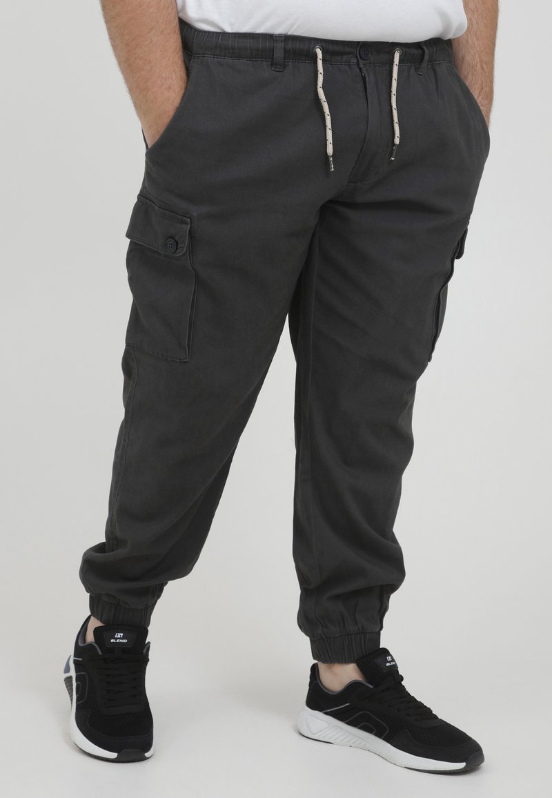 Dunkelgraue Cargo-Hosen mit elastischen Bündchen, seitlichen Taschen und einem verstellbaren Bund. Hergestellt aus einem weichen, strapazierfähigen Stoff. Kombiniert mit schwarzen Sneakers.