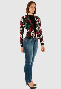 Bloemenpatroon blazer in zwart, groen, crème en rood. Lange mouwen, nauwsluitende pasvorm en open voorkant. Gecombineerd met blauwe jeans en zwarte hakken.