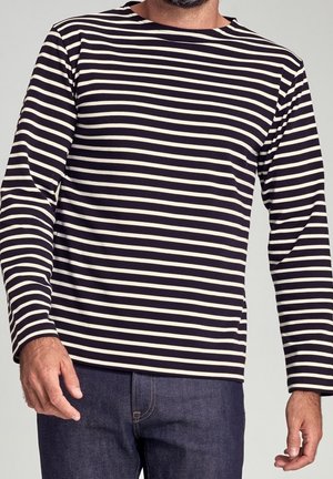 Uomo che indossa una maglia a maniche lunghe con strisce orizzontali bianche e nere e jeans blu scuro, in piedi con le mani rilassate.
