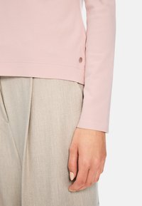 Helles rosa Langarmoberteil aus weichem Material mit abgerundetem Saum, akzentuiert durch ein kleines Logo-Detail unten links. Kombiniert mit beige strukturierten Hosen.