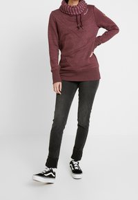 Sudadera de cuello alto en color burdeos con patrones de costura diagonal, puños de canalé y cordones, combinada con jeans oscuros ajustados y zapatillas negras.