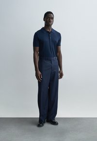 Πόλο μπλε ναυτικό με γιακά, σε συνδυασμό με παντελόνια με λεπτές ρίγες. Τα παντελόνια έχουν εφαρμοστή μέση με ζώνη. Απλά μαύρα παπούτσια ολοκληρώνουν την εμφάνιση.