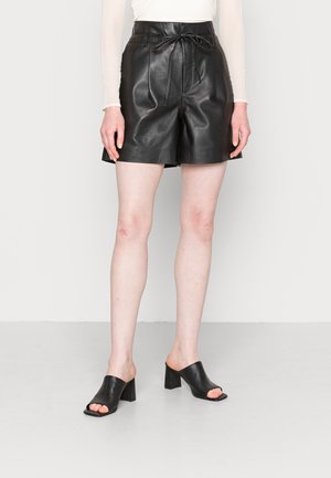Shorts en cuir noir à taille haute, avec un détail à nouer et des jambes larges. Associés avec des mules à talons blocs ouverts noir. Texture lisse sur toute la surface.