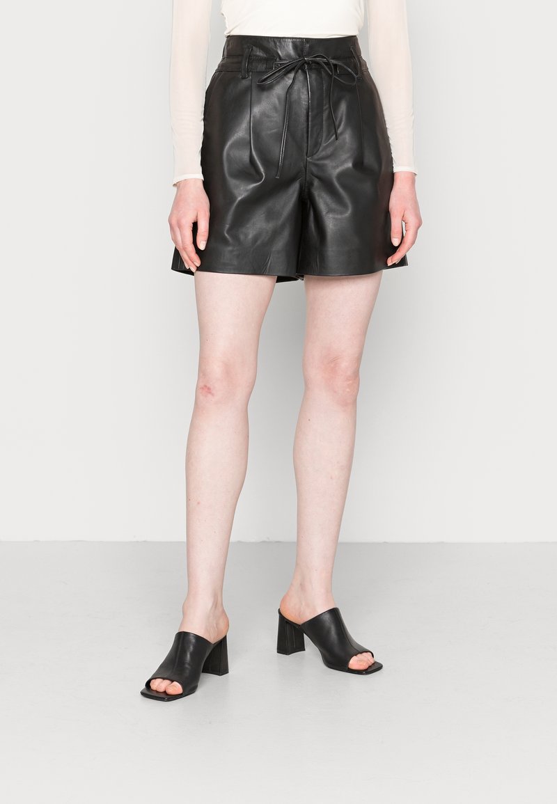 Shorts en cuir noir à taille haute, avec un détail à nouer et des jambes larges. Associés avec des mules à talons blocs ouverts noir. Texture lisse sur toute la surface.