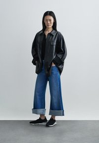 Camisa jaqueta oversized em couro preto, combinada com jeans de pernas largas e bainhas. Os sapatos são pretos, atléticos, com um design elegante.