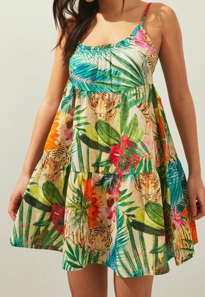 Vrouw met een mouwloze, luchtige jurk met een tropische print van groene bladeren, kleurrijke bloemen en tijgervakken.