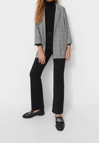 Grijze geruite oversized blazer met brede revers, zwarte coltrui, zwarte uitlopende broek en zwarte loafers met decoratieve metalen accenten.