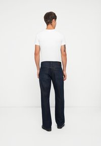 Jeans in denim scuro con una vestibilità rilassata, caratterizzati da un design a gamba dritta e due tasche posteriori. Indossati con una maglietta bianca semplice.