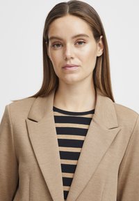 Blazer beige con un diseño ajustado y solapas de muesca, llevado sobre una camisa a rayas negra y beige. Textura suave, silueta entallada, detalles mínimos.
