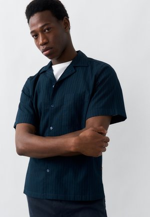 Νέος άνδρας με σταυρωμένα τα χέρια, φορώντας μπλε σκούρο πουκάμισο με κοντά μανίκια και κουμπιά πάνω από λευκό εσώρουχο, στέκεται μπροστά σε απλό ανοιχτόχρωμο φόντο.