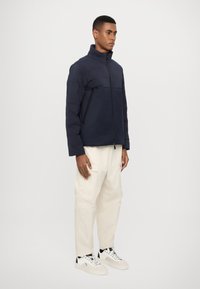 Jachetă puffer de culoare bleumarin cu guler înalt, cu fermoar pe față și design matlasat. Asortată cu pantaloni cargo crem și adidași albi cu accente negre.