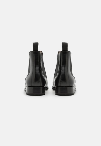 Pareja de botas Chelsea de cuero negro pulido vistas desde atrás, mostrando las lengüetas en el talón y los paneles laterales elásticos sobre fondo blanco.
