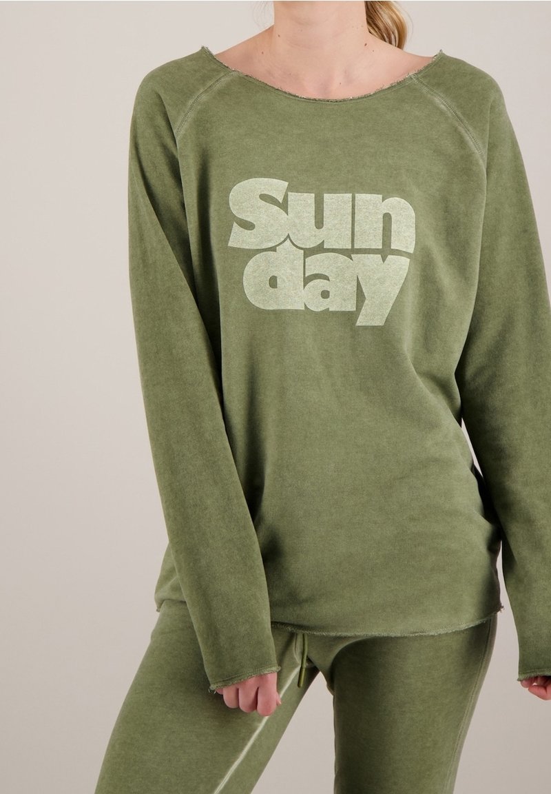 Felpa verde con colletto e polsini usurati, con la scritta "Domenica" stampata in verde chiaro. Abbinata a pantaloni verdi coordinati.