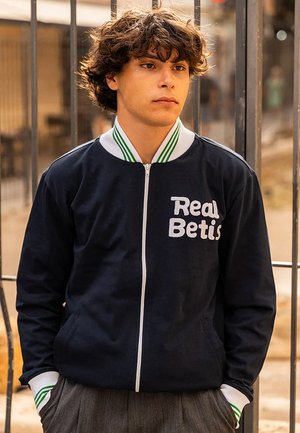 Marineblauwe zip-up jasje met witte tekst "Real Betis". Voorzien van een kraag en manchetten met groene strepen, gemaakt van een zachte, gestructureerde stof en met zijzakken.