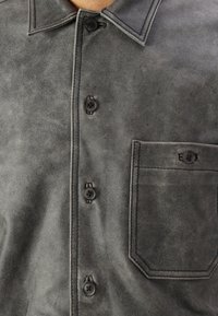Camicia in suede grigio con chiusura a bottoni, colletto a punta e taschino sul petto. Presenta bottoni neri e un tessuto morbido e texture.