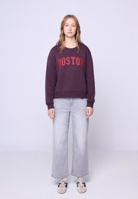 Bluza w ciemnym fioletowym kolorze z napisem "BOSTON 1980" w czerwonych literach, zestawiona z jasnoszarymi spodniami typu wide-leg i szaro-czarnymi sneakersami.