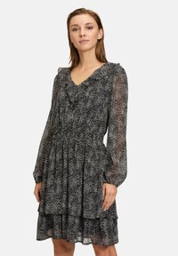 Zwart-wit polkadotjurk met ruches bij de halslijn, lange doorschijnende mouwen en een gelaagde rok. Gemaakt van lichtgewicht, vloeiend materiaal.