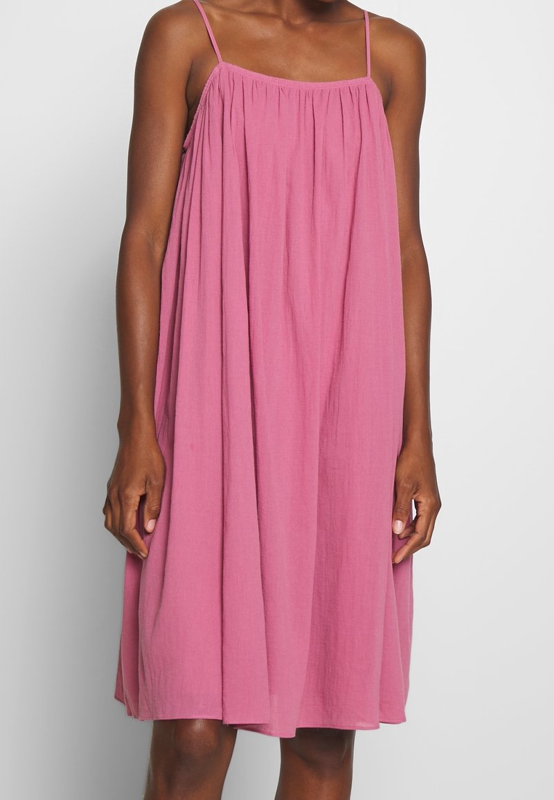 Femme portant une robe ample rose sans manches, longueur genoux, avec de fines bretelles, posant devant un fond uni clair.