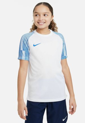 Ein lächelndes Kind trägt ein weißes Nike-Shirt mit blauen gestreiften Ärmeln, kombiniert mit dunklen Shorts, vor einem neutralen Hintergrund.