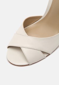 Sandale à talons hauts en cuir blanc avec un design à lanières croisées, bout ouvert, texture lisse et un subtil accent de couture le long des bords.