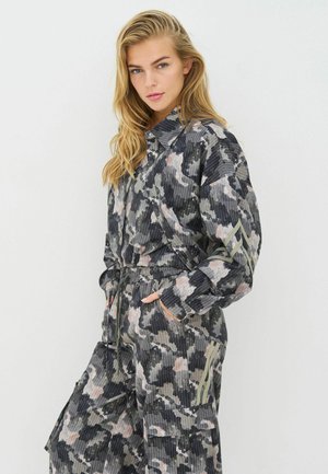 Maskuojamosios raštų kombinezonas pilkos, žalios ir rožinės spalvų. Turi apykaklę, elastinę liemens dalį ir kišenes. Audinys su tekstūra ir vertikaliomis juostelėmis.