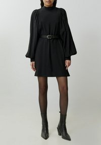 Zwarte jurk met hoge kraag en lange pofmouwen, strak getrokken in de taille met een zwarte riem. Draag het met zwarte enkellaarsjes met hak.