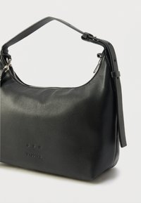 Geantă de mână din piele neagră cu formă rotunjită, prevăzută cu închidere superioară cu fermoar și o curea scurtă, reglabilă. Logo-ul este gravat pe partea din față.