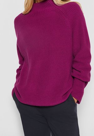 Femme portant un pull violet à manches longues, côtelé, avec un col montant, et un pantalon noir, les mains dans les poches, sur un fond uni.