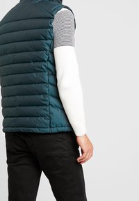 Teal pufferväst med horisontell vaddering, bärs över en vit stickad tröja med ett texturerat grått mönster. Svarta byxor syns nederst.