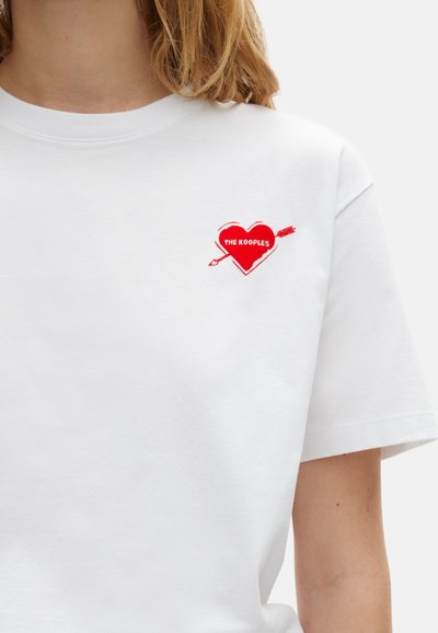 Hvid kortærmet t-shirt med en rød hjertegrafik og en pil samt teksten "THE KOOPLES" i fed skrift, hvilket skaber en slående kontrast.