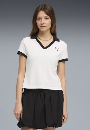 Giovane donna con capelli lunghi castani che indossa una camicia bianca a maniche corte con colletto e rifiniture nere, e una gonna nera, su sfondo grigio.