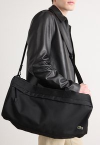 Bolsa de tote negra hecha de un tejido duradero, con una forma rectangular elegante, dos asas superiores y un logotipo bordado en el lateral.