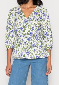 Blouse à fleurs sur fond blanc, avec des fleurs bleues et des feuilles vertes. Elle possède un col en V, des manches sept huitièmes et un détail peplum.