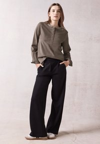 Camicia verde oliva a maniche lunghe con dettagli di bottoni, abbinata a pantaloni neri larghi e sneakers bianche. Texture del tessuto lisce visibili.