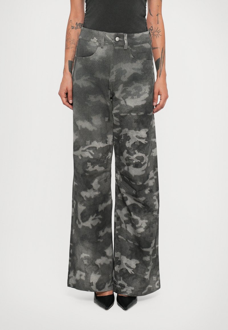 Personne portant un pantalon en camouflage gris taille haute et large avec des chaussures noires à bout pointu, se tenant avec les bras détendus le long du corps.