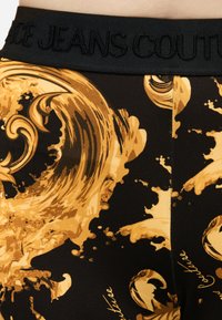 Tela negra con patrón ornamental barroco en dorado y cinturilla negra con la etiqueta "JEANS COUTURE" en relieve.