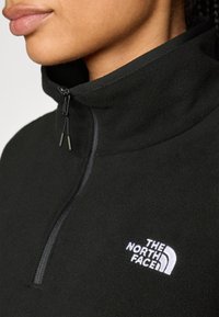 Svart fleece-pullover med høy krage og halv glidelås, med en hvit brodert logo på brystet og en teksturert stoffoverflate.