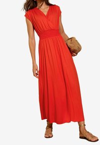 Abito maxi rosso senza maniche con scollatura a V e vita arricciata. Tessuto fluido e design lungo fino alla caviglia. Indossato con sandali dorati e una borsa intrecciata.