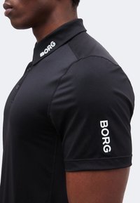 Κοντινό πλάνο ενός ατόμου που φοράει μαύρο κοντομάνικο πόλο μπλουζάκι με λευκό κείμενο "BORG" στο γιακά και το μανίκι.