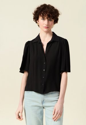 Frau mit kurzen lockigen Haaren, die goldene Ohrringe, eine schwarze kurzärmelige Bluse mit Knöpfen und hellblaue Jeans trägt, steht vor einem einfarbigen Hintergrund.