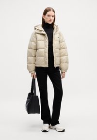 Bežs pūkains jaka ar kapuci, izšūšanas dizains, ar rāvējslēdzēja aizdari, kopā ar melnu turtleneck, melnām biksēm un melnu rokassomu.