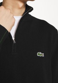 Svart stickad tröja med dragkedja vid halsen, med en liten grön krokodil-logga. Den har en slät textur och en kontrasterande grön sömdekoration.