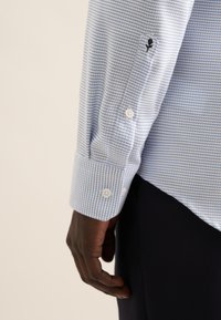 Camicia button-up azzurro chiaro con un motivo a piccole scaglie, caratterizzata da bottoni bianchi e un ricamo di rosa nera sul polsino. Texture liscia.