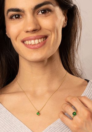 Gold chain necklace with a green gemstone pendant, matching green gemstone ring, worn by a woman with dark hair. Light gray top.